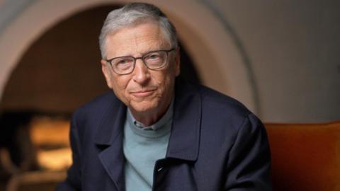 Bill Gates in blue jumper and jacket, sitting by fireplace