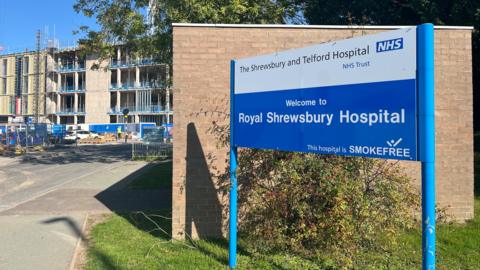 There is a large blue and white sign at a walled entrance with the words "Royal Shrewsbury Hospital" on it. The sign is set on a grassed area with a road running alongside. There is a partly-built hospital unit behind.