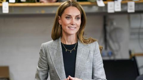 Catherine, Princess of Wales with long brown hair is wearing a grey and black blazer and black top and a gold necklace with the letters 'G, C, L' on it with small gold hoop earrings. She is smiling to the side with her hands clasped together. The background is blurred.
