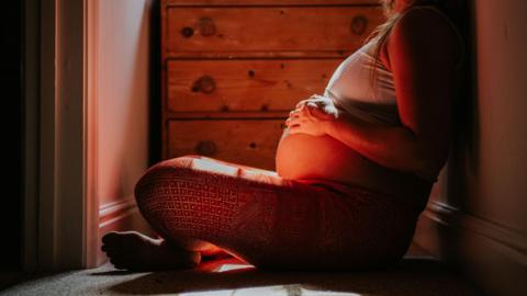 A woman sits on the floor with her hands on her pregnant belly. There is a chest of drawers in the background. 