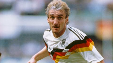 West Germany striker Rudi Voller captured mid‑stride during a match. He wears the unmistakable white kit with the bold diagonal black‑red‑gold sash across the chest — one of the most iconic designs in international football. The team crest sits on the left side of the shirt. His expression is concentrated, eyes fixed ahead, with light blond hair and a distinct moustache. The background is blurred stadium colour — crowd, pitch, movement 