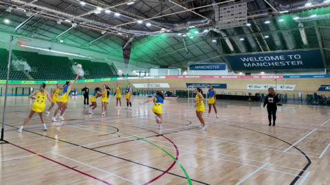 Manchester Thunder players on the netball court wearing yellow sports dresses. Half of them wear blue bibs. One player is jumping to block the ball.