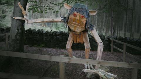 A wooden sculpture of the Buggane, which has long dark hair with its ear sticking out, red eyes and protruding teeth, along with long arms.