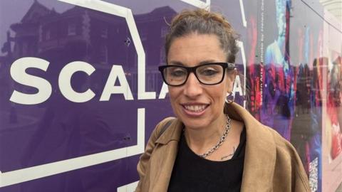 A woman with brown hair tied in a bun, wearing black glasses, silver hoop earrings, a silver necklace and a black top with a beige jacket over it. She is standing in front of a board that is purple, in white writing it reads "Scala"