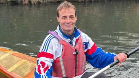 A Caucasian man sitting inside an orange kayak boat holding a black and blue paddle stick. The boat is in the canal. The man is wearing a Team GB jacket that is white, red and blue. He is also wearing a red life jacket. 