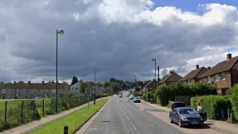 A main road on a quiet housing estate. There are cars parked on the right and playing fields on the left.