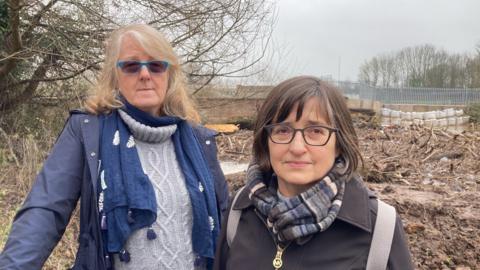 Two women stood at a river side. The woman on the left is wearing a blue coat with blue scarf and grey knitted top. She has blonde hair and is wearing blue-rimmed glasses. Next to her is a woman with brown hair and brown glasses. She is wearing a brown coat with a grey and blue patterned scarf