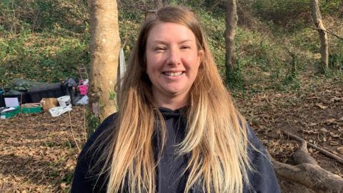 Youth Commission for Guernsey and Alderney senior youth worker Cleo Thomas smiles while stood in a forest area. She has long straight hair which reaches down to her chest. She has a pair of glasses propped on top of her head.