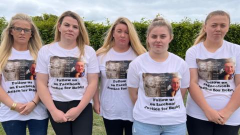 Five sisters, side-by-side. There are all looking to the camera. They are all wearing jeans and have the same white T-shirt on. It states "Follow us on Facebook. Justice for Bobby". They are standing on grass and there is a green hedge behind them. 