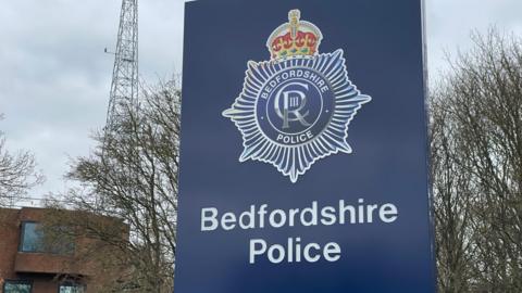 A close up shot of the large blue sign outside Bedfordshire Police headquarters. It has the police crest on and the words Bedfordshire Police in white font. There is a red brick building to the left, and bare trees behind.