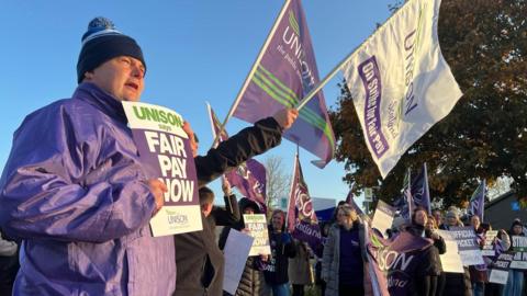 People stand on a picket line at a Perth school, the man in the foreground is wearing a Unison bobble hat in purple and green holding a placard saying "fair pay now", with a line of other strikers stretching from left to right behind him