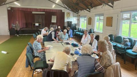 The women are sitting around two tables on the right-hand side of the hall. In the background is the stage where The Beatles once played. On the left, a game of boules is being set up.