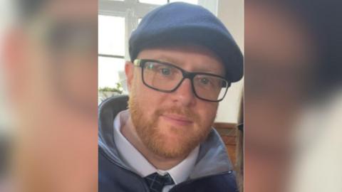 A man in his late 30s, with short, ginger beard and thick-rimmed black glasses. He is wearing a collar and tie under a jacket.