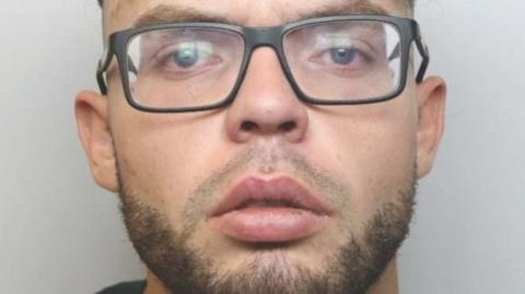 A mugshot of a young man with black-framed glasses and a short beard. 