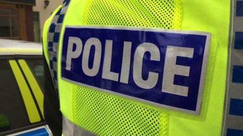 A police officer standing in front of a police car. The image shows a close up of the word "Police" printed on the back of their vest.