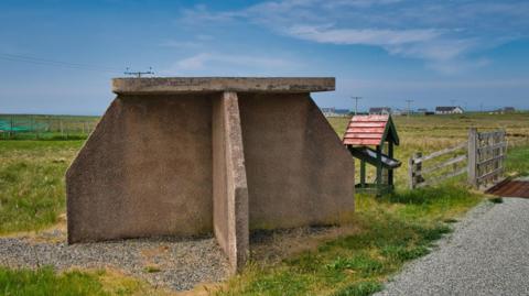 A bus stop in the Western Isles. It is made up of a number of shaped concrete slabs, with one on top as a roof. The bus stop is on a rural road next. It is a sunny day.