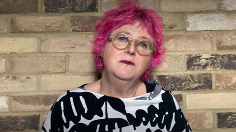A woman with short pink hair, wearing glasses and a black and cream top. There is a brick wall behind her.