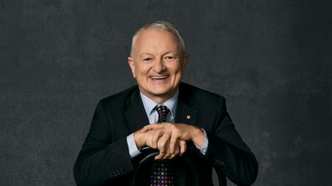 The ABC's chief elections analyst, Antony Green, smiles broadly at the camera as he sits backwards on a chair. 