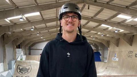 Jayden Sharman smiling into the camera at Adrenaline Alley skatepark. He has a grey helmet on, round framed glasses and a black hoodie. Large wooden ramps are visible in the background.