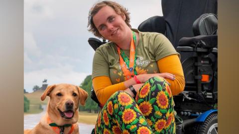 A woman in an orange and green top and trousers with sunflowers on. She is sitting in front of her wheelchair and has assistance dog Ocho sat on the ground next to her.