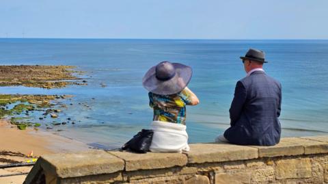 A photohraph of a woman and a man sitting on a wall looking out towards the sea 