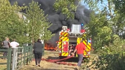 A fire engine with a long red hose coming out of the back is parked in front of large dark grey plumes of smoke, with orange flames visible. The setting is rural, with several trees nearby and a large building just visible behind trees near the fire.