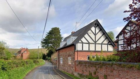 A cottage stands near a road with a walled garden. The walls are partly brick and partly black and white. Across the road, there is a grassy verge and hedge. Further cottages are around the property. There is open countryside further away.
