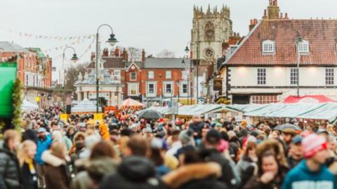 Crowds of people in the centre of Beverley with market stalls, buildings and lights and bunting in the background