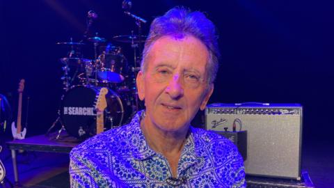 Frank Allen, is looking at the camera. He is in front of a drum kit and is wearing a patterned shirt.