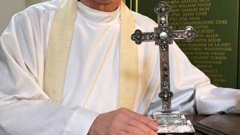 A close up picturing showing the Reverend David Minns holding the base of the silver cross, which is sitting on top of a stone altar. The cross is intricately engraved with a flowery and dotted pattern.