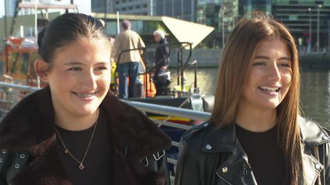 Twins Lola and Evie, both wearing dark jackets over dark tops, seen at Salford Quays with an orange RNLI lifeboat in the background