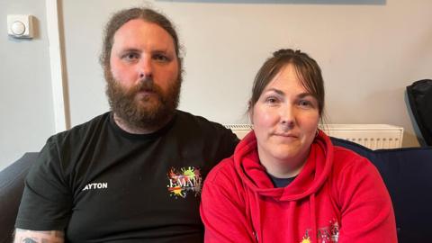 Layton Jones (left) with partner Gemma Baker (right), they both sit on a sofa. Gemma wears a red pull over hoodie and looks at the camera. She has brown hair with sweeping bangs and the rest pulled back. Mr Jones has a long light brown beard and long hair parted in the middle tied back. 
