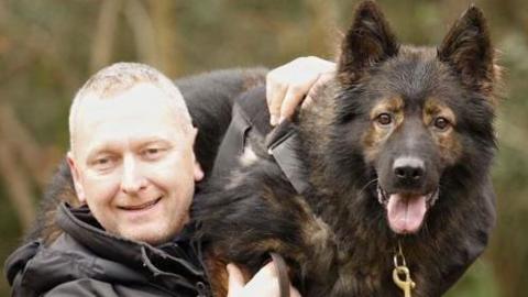 A blond man in a black jacket holding a Belgian Malinois on his left shoulder. The dog has a black lead with gold chain and its tongue is hanging out. The man is smiling. They are standing outside, with greenery out of focus behind.