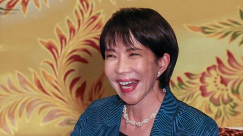 Sanae Takaichi smiling wearing a blue suit against a yellow background