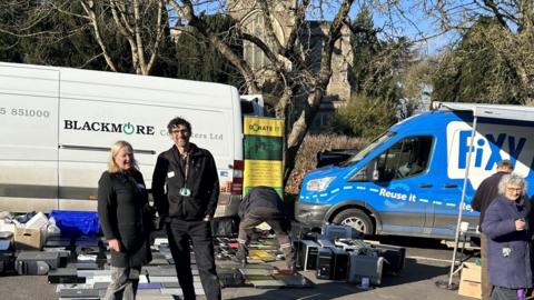 Two people dressed in black and grey clothing standing in front of two vans. A variety of devices cover the ground behind them.