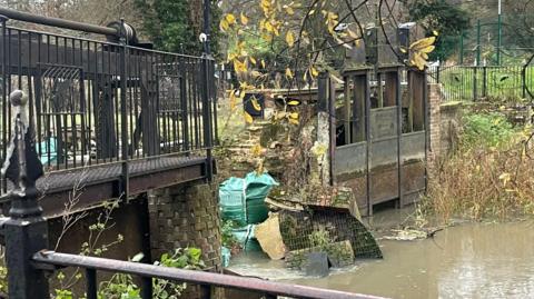 The weir in its collapsed state. There is brickwork that has collapsed into the water. This brickwork was once the top of the weir and its side. There is a green sandbag is also in the river. The parts still in place are a mixture of wooden and iron structures.
