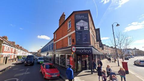 The outside of Morleys Tooting department store, showing signs with black backgrounds and white words reading "Morleys" on the front. There are people on the street around the store and you can see rows of houses on either side.