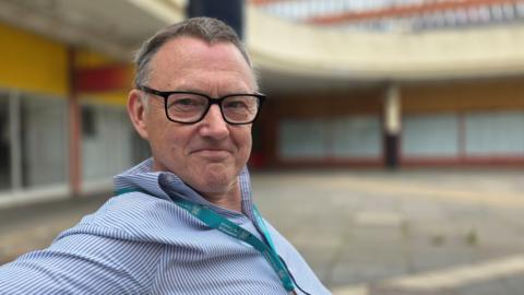 Mike Stonard is sitting on a bench in Anglia Square. He's wearing a blue and white pinstripe shirt along with a light blue lanyard. He also is wearing glasses.