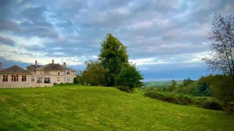 High Borrans, a large cream-coloured stately home on a grassy ridge overlooks rolling hills and forests to the right. The sky above is gloomy and dramatic with dark clouds.
