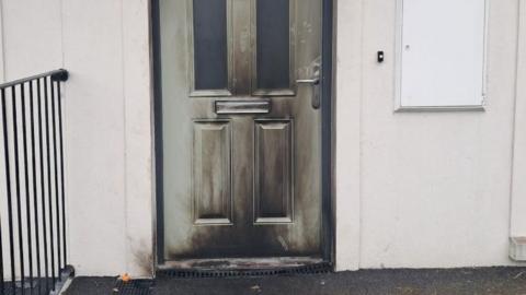 A green door with black fire marks over most of it. There is a large number in the middle of the door. The walls of the house are cream.