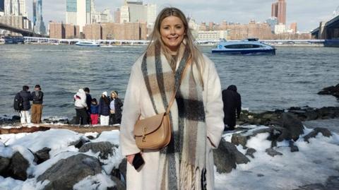 Faye Morton, a woman with long blonde hair wearing a long cream coat with a brown, black and cream-checked scarf and a brown handbag. She is smiling and standing on a snowy harbour with the sea and tower blocks in the background. 