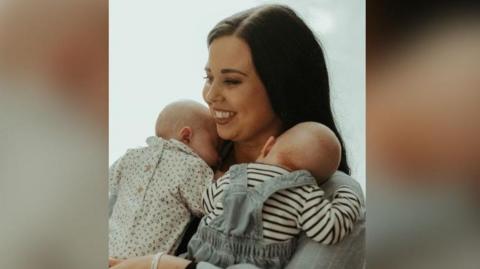 The photo shows a woman with brown hair holding two newborn babies. She is smiling and is looking to the left of the photo. One baby is wearing a flowery baby grow, and the other is wearing dungarees and a striped top. They both have their heads on the woman's shoulders. 