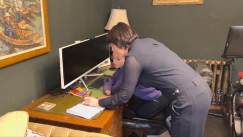 a mum and daughter lean over a notebook set on a wooden desk with a white computer monitor in front of them