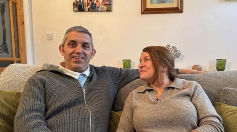 Phill Panter wearing a grey jumper sat on a grey sofa facing the camera, with his wife, Yvonne, facing him