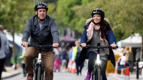 A man and woman ride their bikes, wearing helmets and coats. They're cycling down a road with a blurred, busy market in the background. 