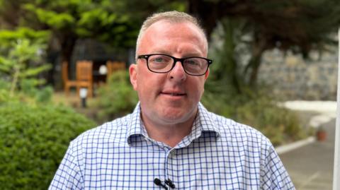 A headshot of a Garin Dart, a man with short brown hair and black glasses. He is wearing a black and blue checked shirt.