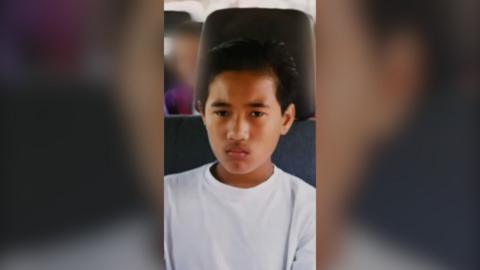 A young boy with short black hair sits in a motor vehicle. He is wearing a white T-shirt and looks upset.