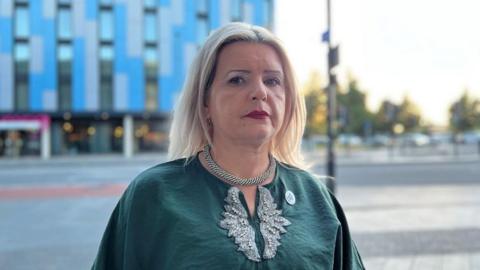 Tatiana is wearing a silver chain and a green top and is behind Slough high street. 