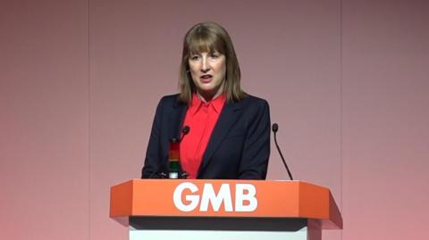 Reeves at a podium with the letters GMB on it. She is wearing a dark jacket and a red shirt.