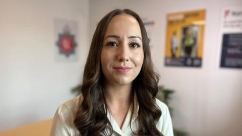 Jo Webb wearing a light coloured shirt with long brown hair smiling at the camera. She is inside a white room with posters and the background is out of focus.